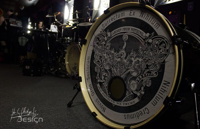 The bass drum head, lit by whatever was left of the house rig. The engraved crest — Surrectum Ex Nihilum, Novus Ordo Seclorum, Nihilum Credimus, In Nothing We Trust — is the band's own artwork. I only photographed it. [wide]